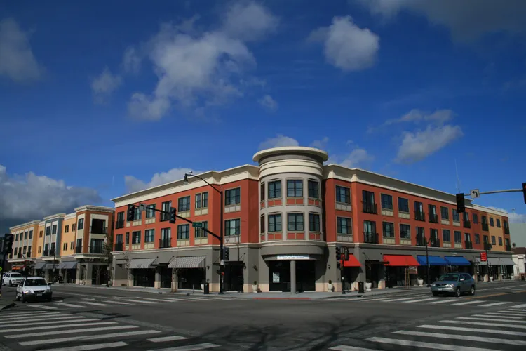 A modern building with large glass windows at a street intersection, featuring storefronts and multiple floors under a blue sky.