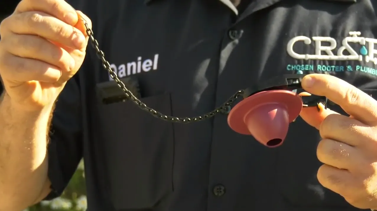 A person in a Chosen Rooter & Plumbing, Inc. uniform holding a toilet flapper chain and valve outdoors. A person in a Chosen Rooter & Plumbing, Inc. uniform holding a toilet flapper chain and valve outdoors.