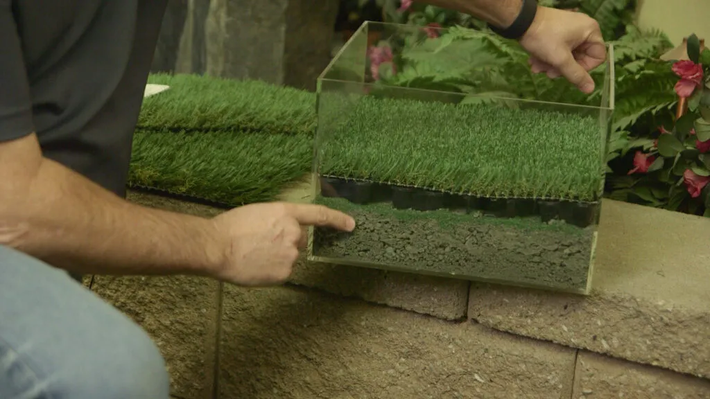  A person points at a sample box showing layers of artificial turf, soil, and base. Artificial grass samples lie in the background, with plants visible nearby.