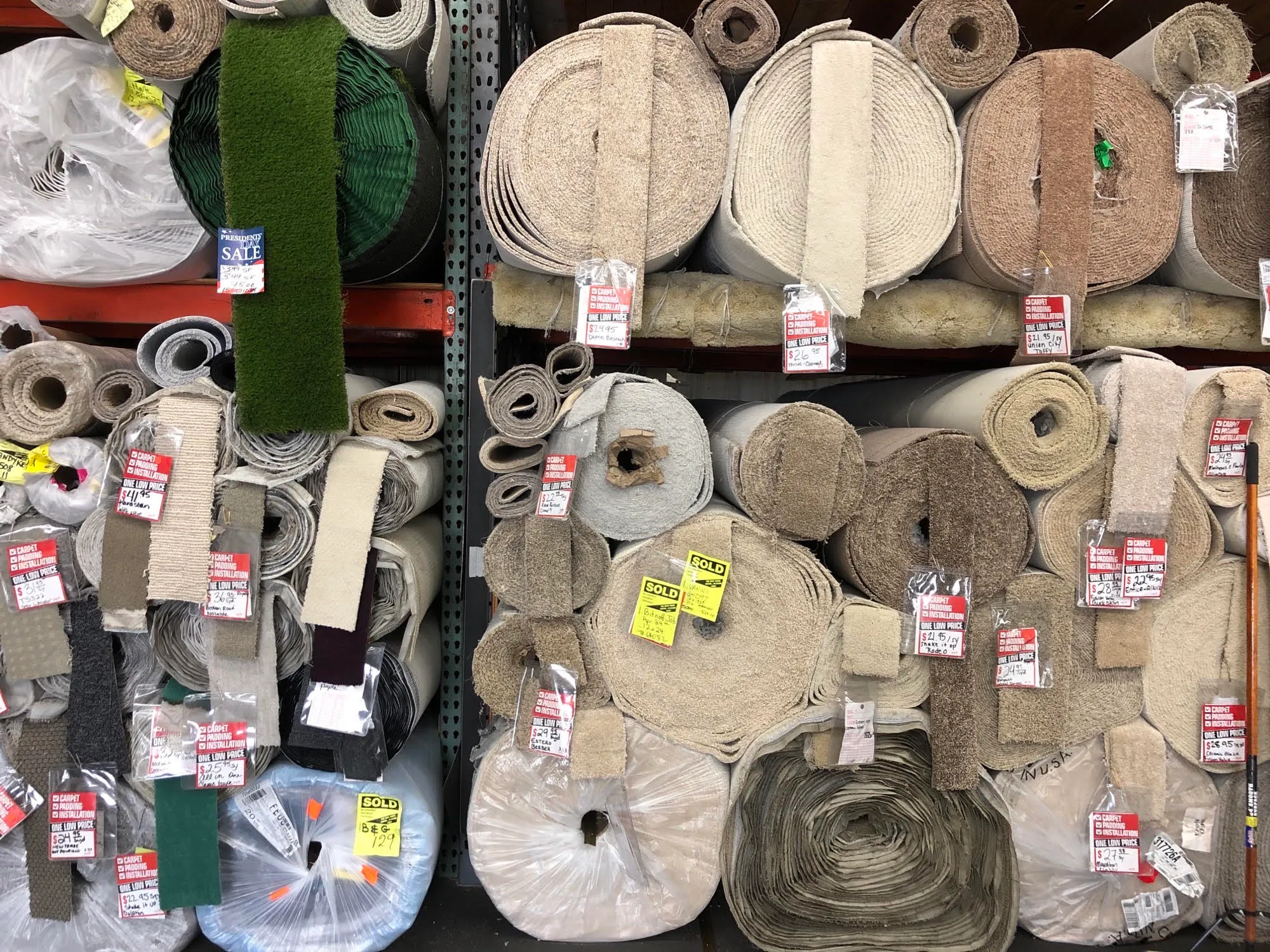  Various carpet rolls in different colors and textures stacked on shelves, each marked with sale tags and prices.