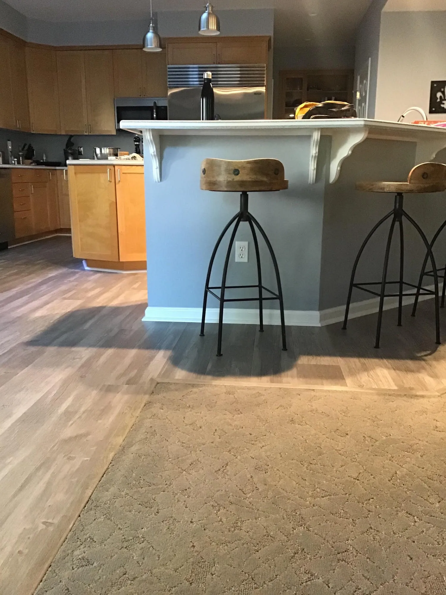  A kitchen with wooden cabinets, metal bar stools, and a countertop. The floor transitions from carpet to hardwood.