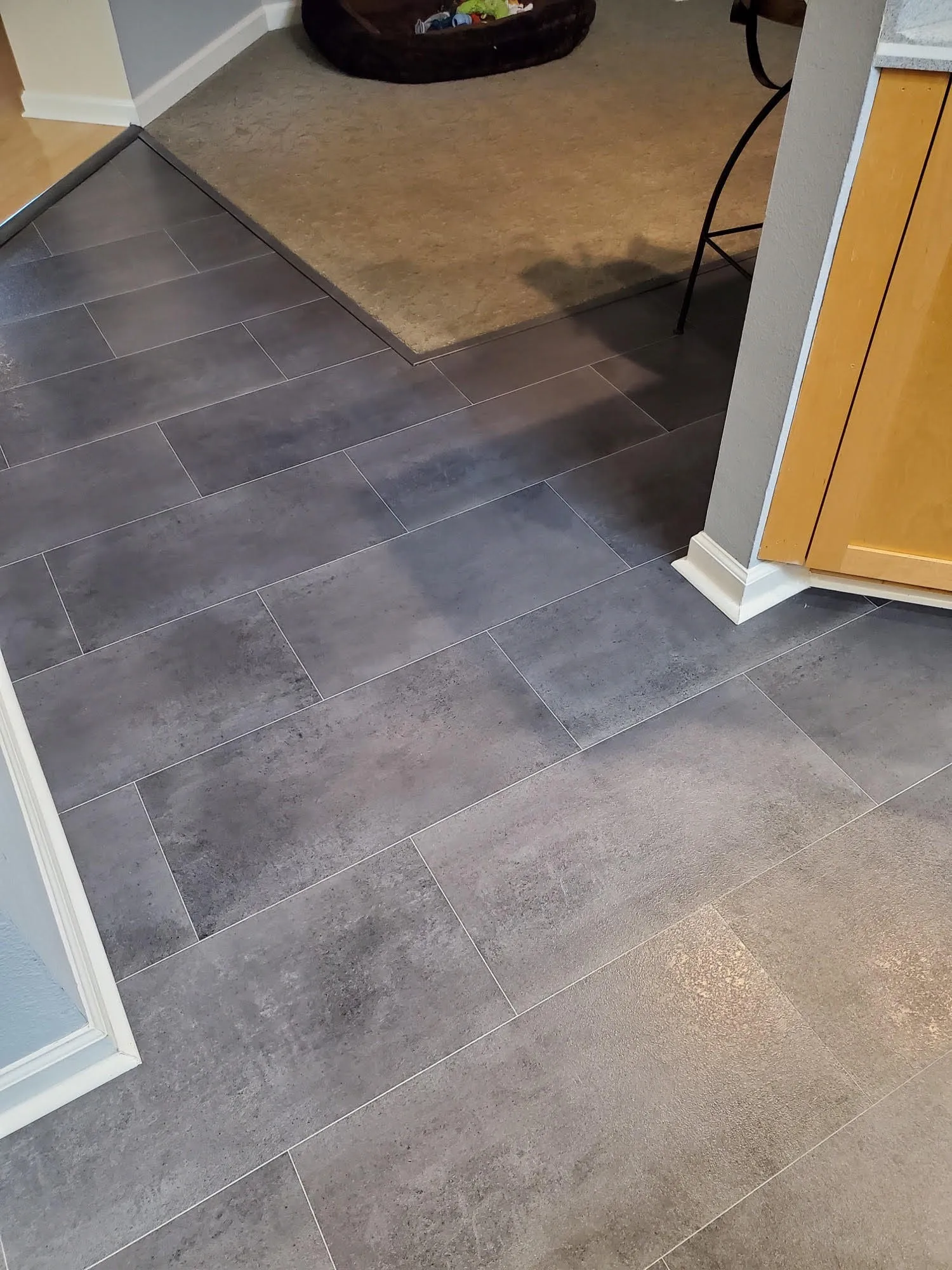  Gray tile flooring transitions to carpet in a residential setting. Nearby are a wooden cabinet and a chair.