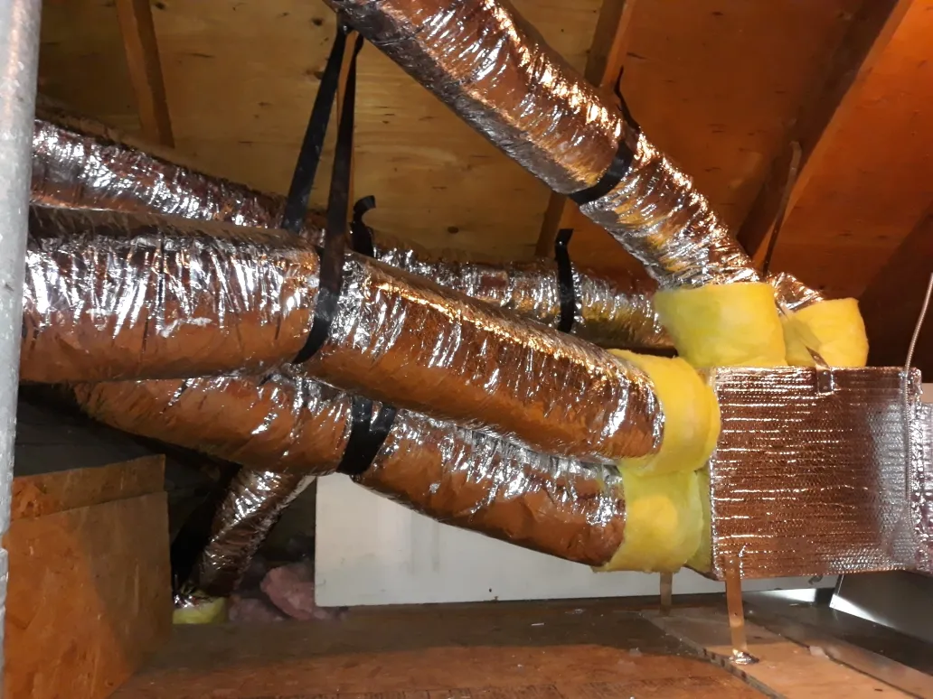 Insulated ductwork in an attic, held up by straps, with a reflective surface and yellow insulation wrapping around sections. Insulated ductwork in an attic, held up by straps, with a reflective surface and yellow insulation wrapping around sections.