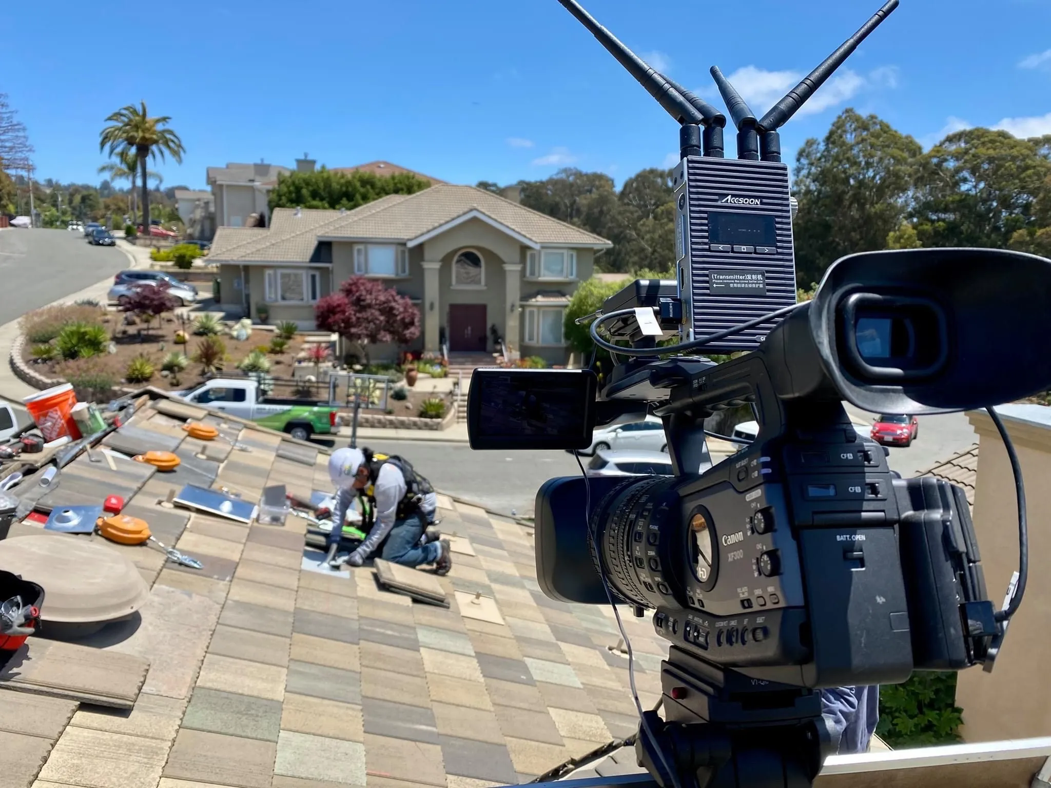  Rooftop worker installing solar panels; camera in foreground. Residential street and houses visible under clear sky.