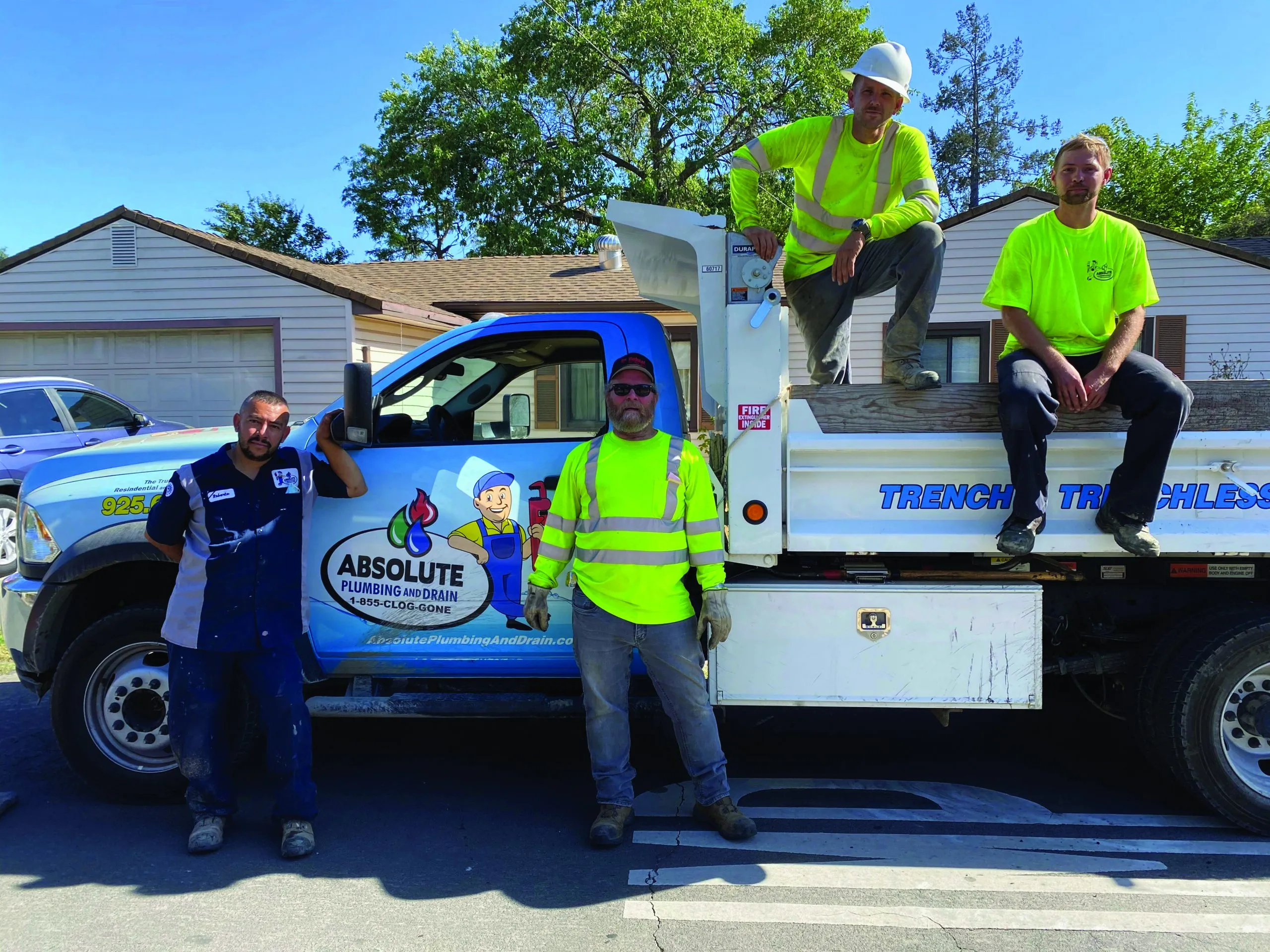Four people in neon safety gear and work uniforms stand and sit on a plumbing truck labeled "Absolute Plumbing and Drain" on a suburban street. Four people in neon safety gear and work uniforms stand and sit on a plumbing truck labeled "Absolute Plumbing and Drain" on a suburban street.