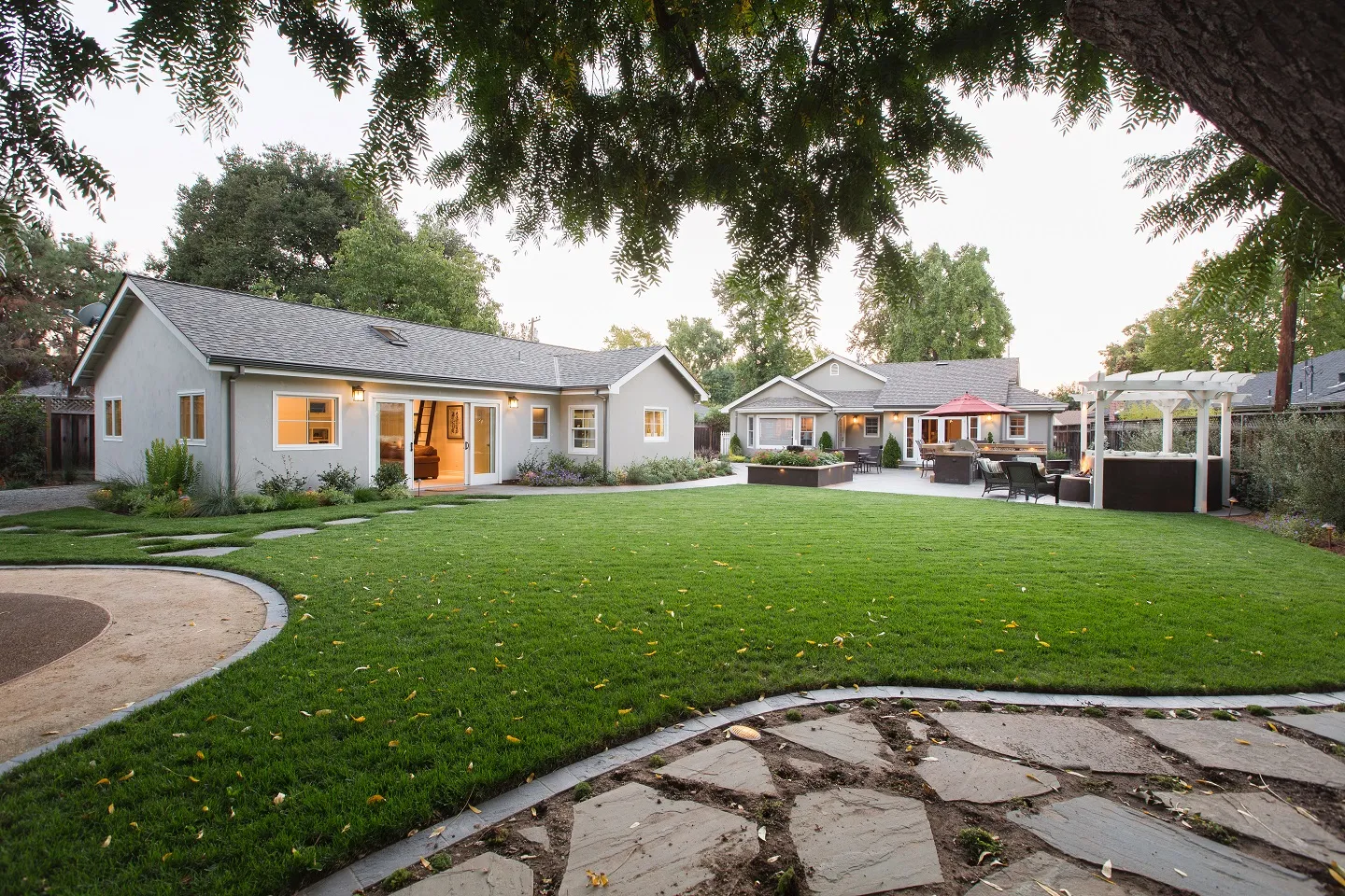 A landscaped backyard with a green lawn, patio, outdoor seating, and a detached house in the background.