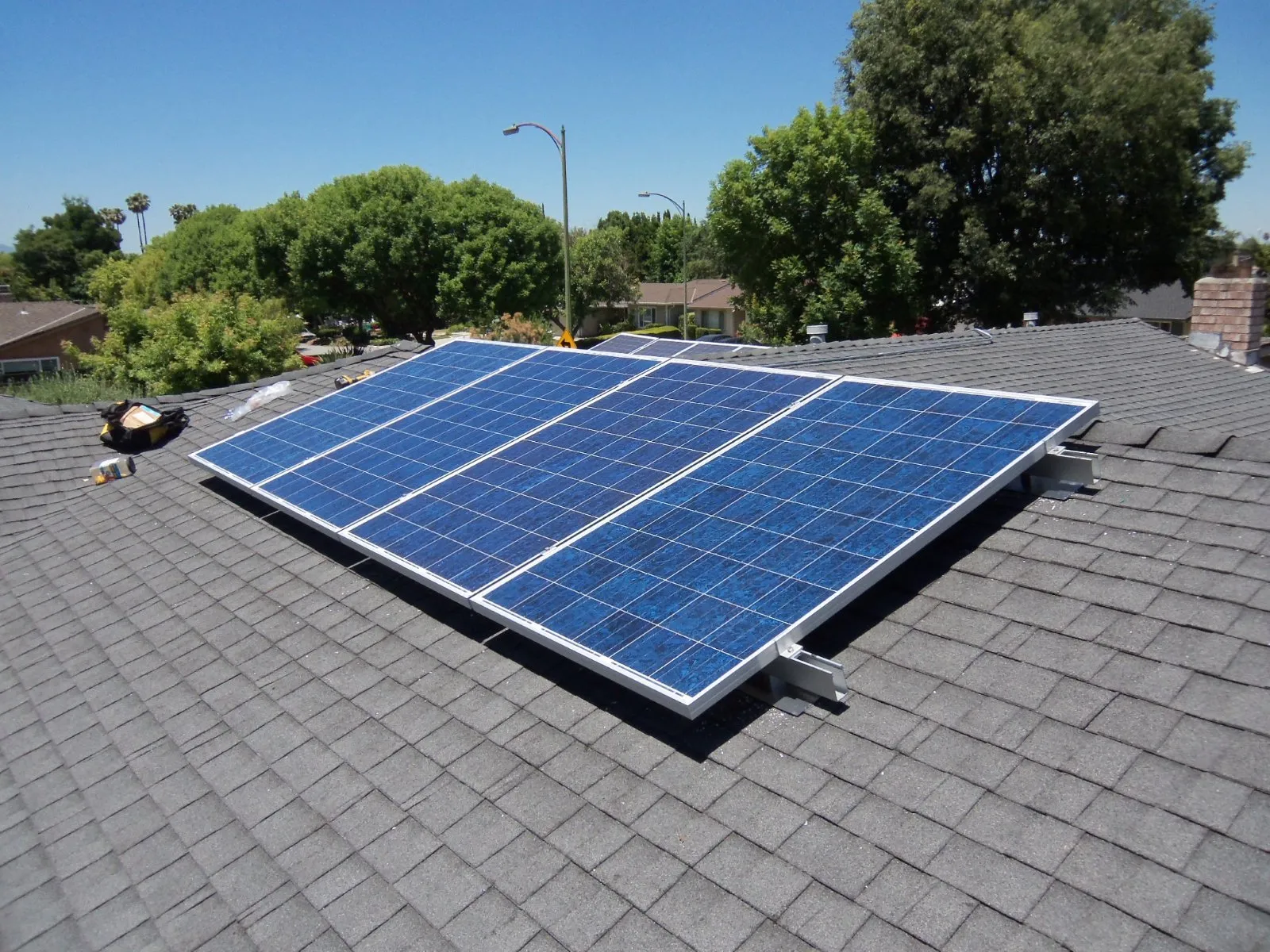  Rooftop with solar panels, surrounded by trees, under a clear blue sky. Tools and a bag are on the roof nearby.