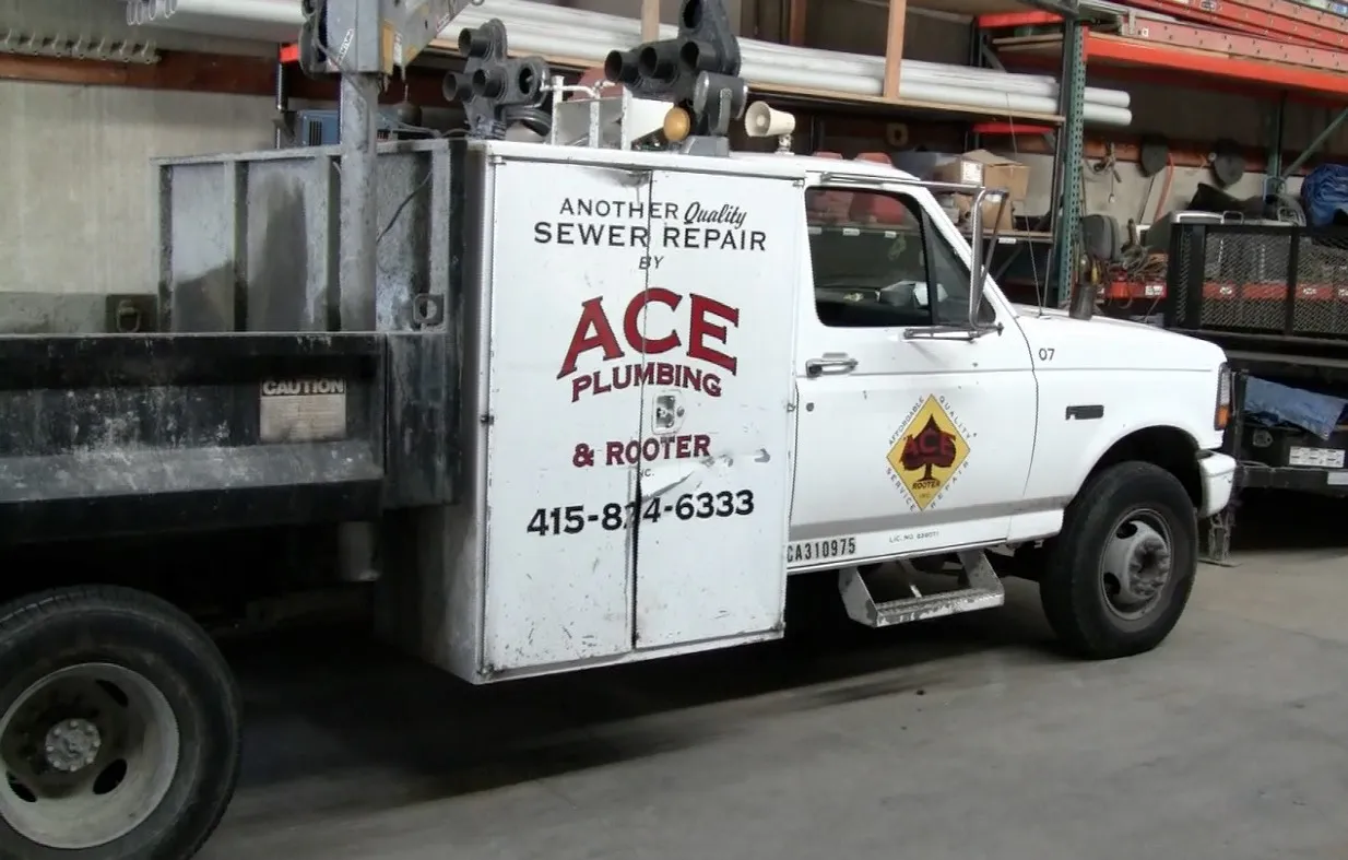 An Ace Plumbing and Rooter truck inside a workshop with various tools and materials stored on shelves. The truck's side displays the company logo and contact information. An Ace Plumbing and Rooter truck inside a workshop with various tools and materials stored on shelves. The truck's side displays the company logo and contact information.