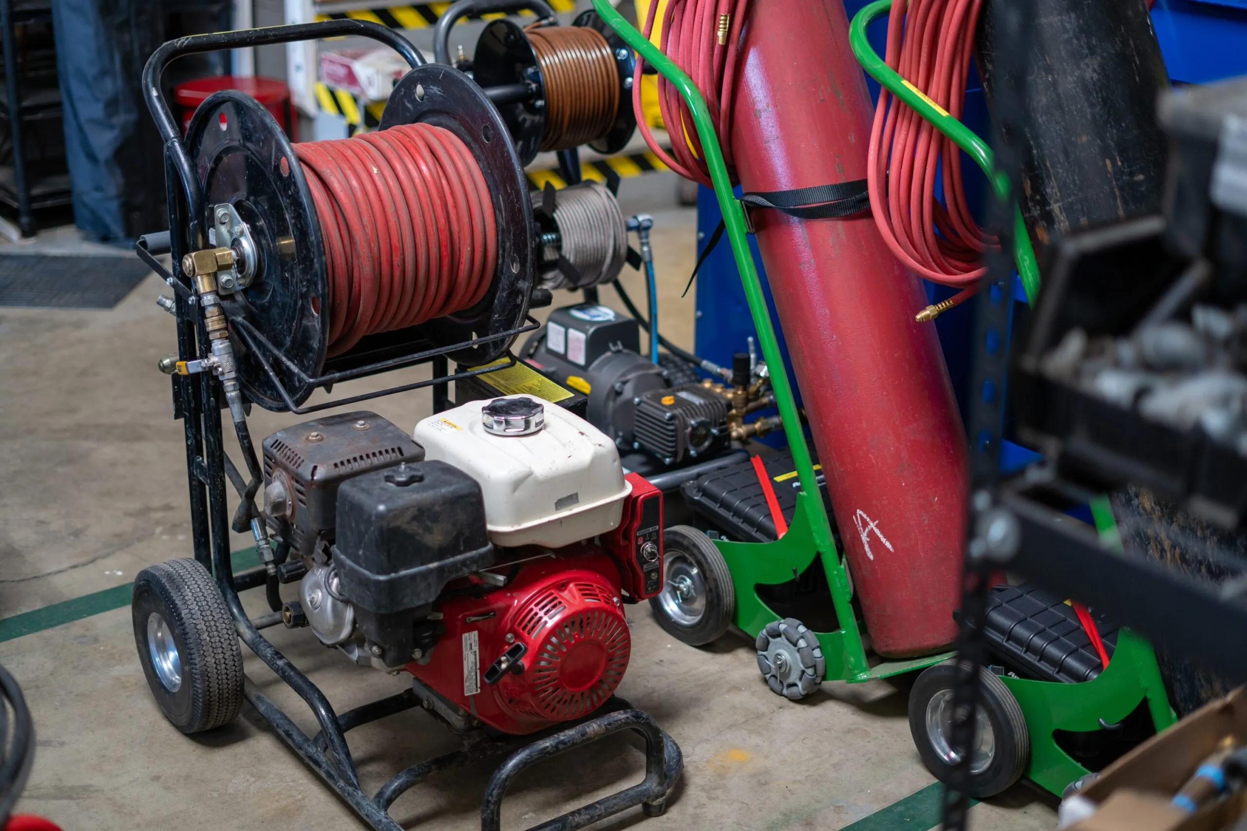 Close-up of hose reels and machinery in a workshop, including a red hose, engine, and various tools on a concrete floor. Close-up of hose reels and machinery in a workshop, including a red hose, engine, and various tools on a concrete floor.