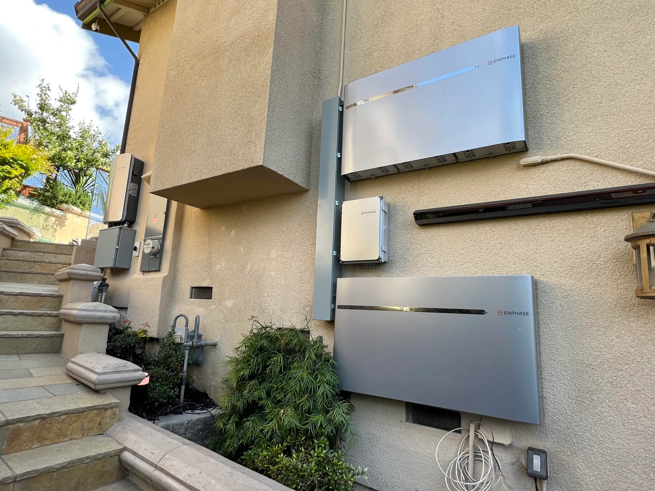  Solar battery systems mounted on the exterior wall of a building, with electrical boxes and wiring visible. Stone steps and plants are nearby.