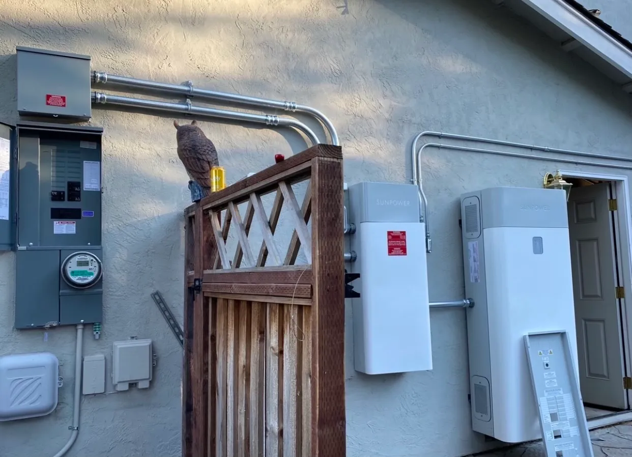  Electrical panels and solar equipment installed on the exterior wall of a building, with conduit pipes and a wooden fence nearby.