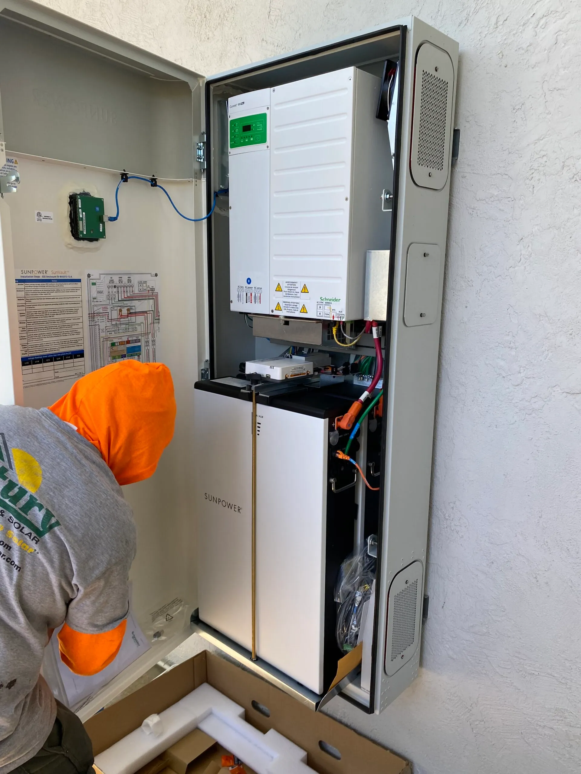  Technician in an orange hooded sweatshirt installing an energy system labeled SunPower inside an open electrical panel.