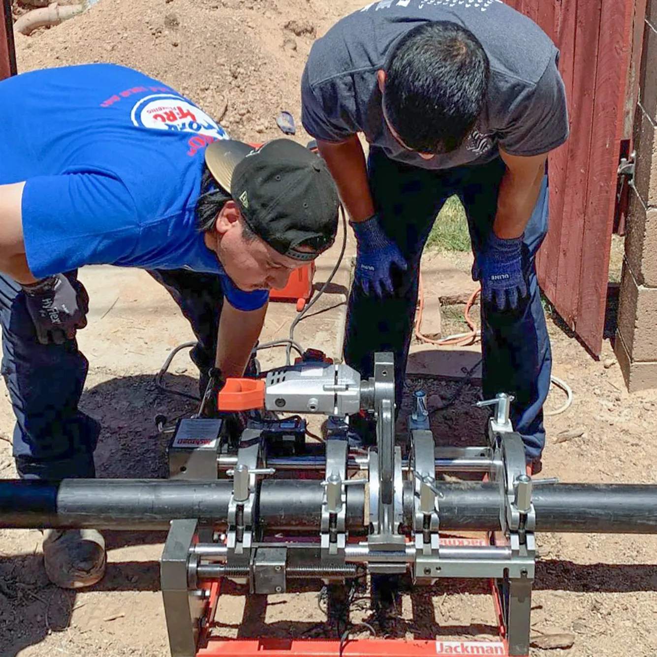 Two technicians using a pipe fusion machine outdoors. One is in a blue shirt and hat, the other in a gray shirt and gloves. A large black pipe is clamped in the machine.