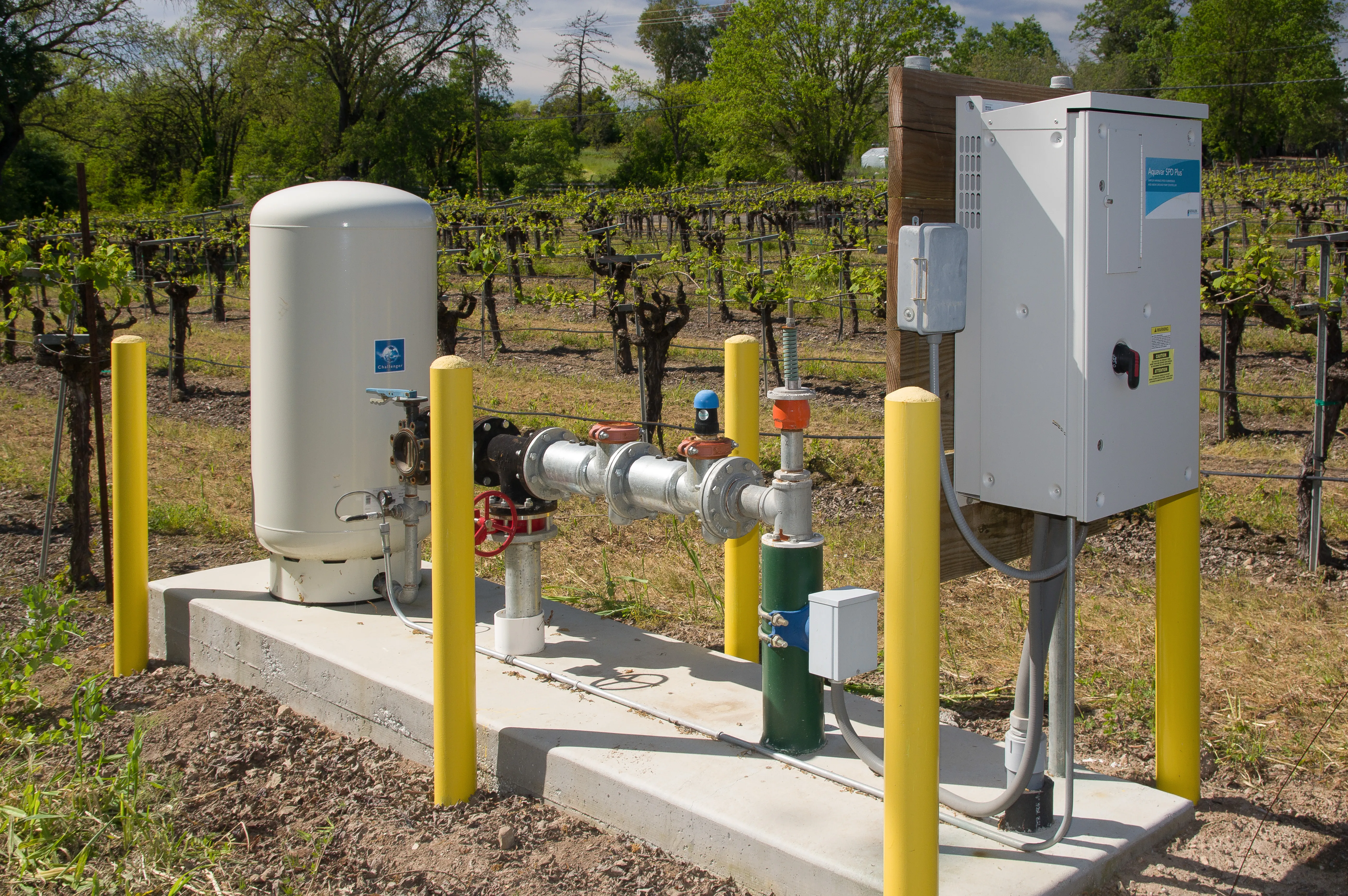 Water pump system with pipes, valves, and control panel, set on concrete slab in a vineyard. Surrounded by yellow posts and grapevines in the background.