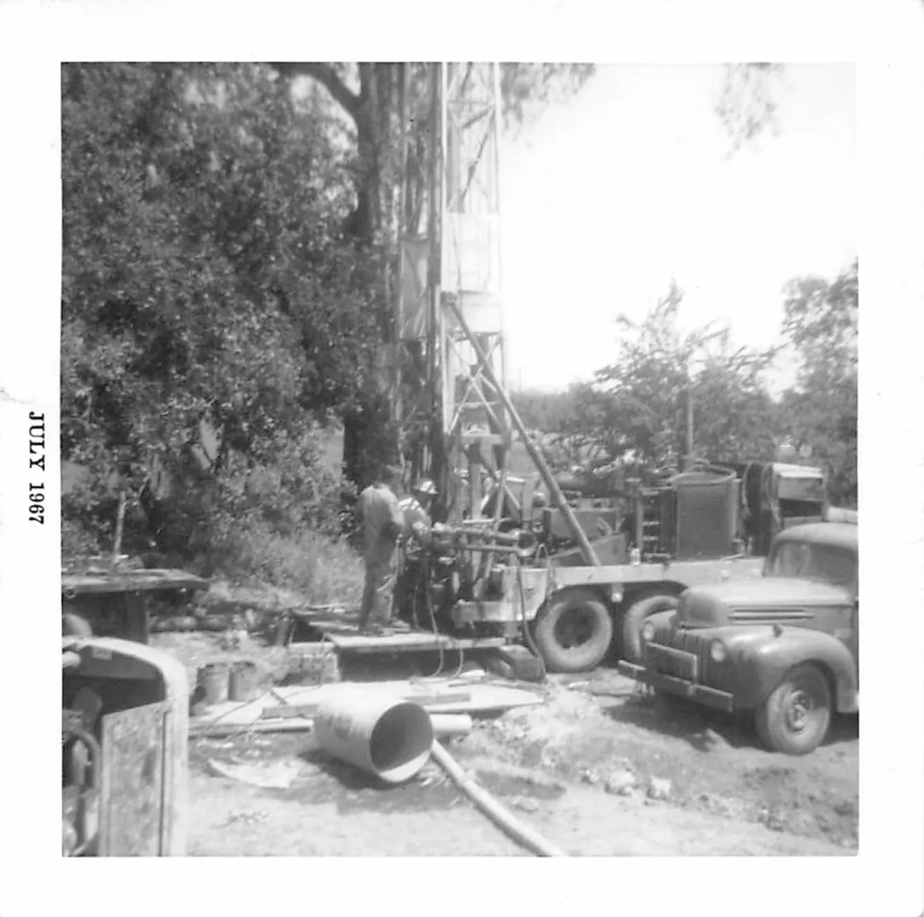  A 1967 scene showing a worker operating drilling equipment beside an old truck, surrounded by trees and scattered tools.