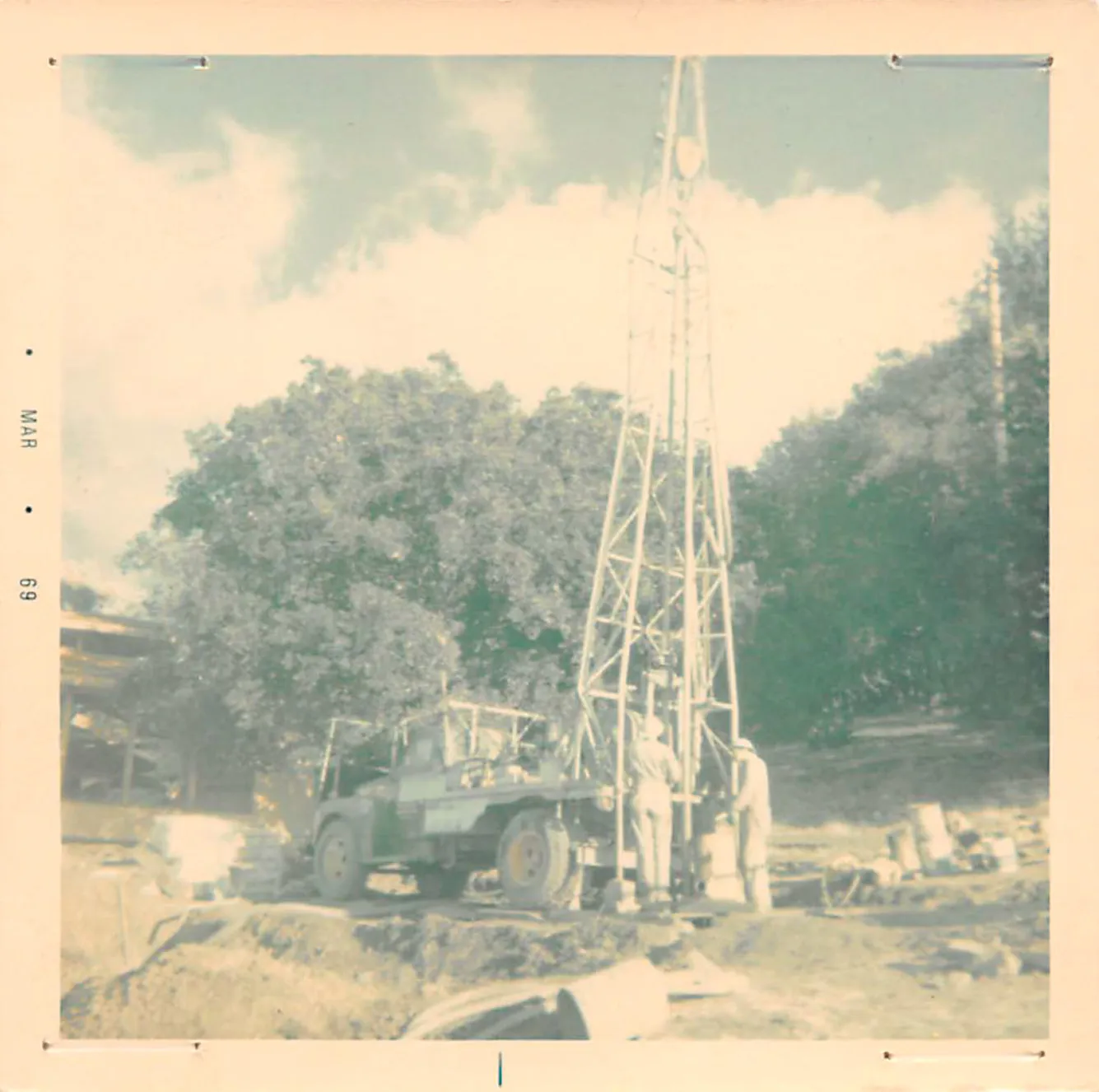  Workers operate a drilling rig next to a truck in a rural setting with trees, under a partly cloudy sky.
