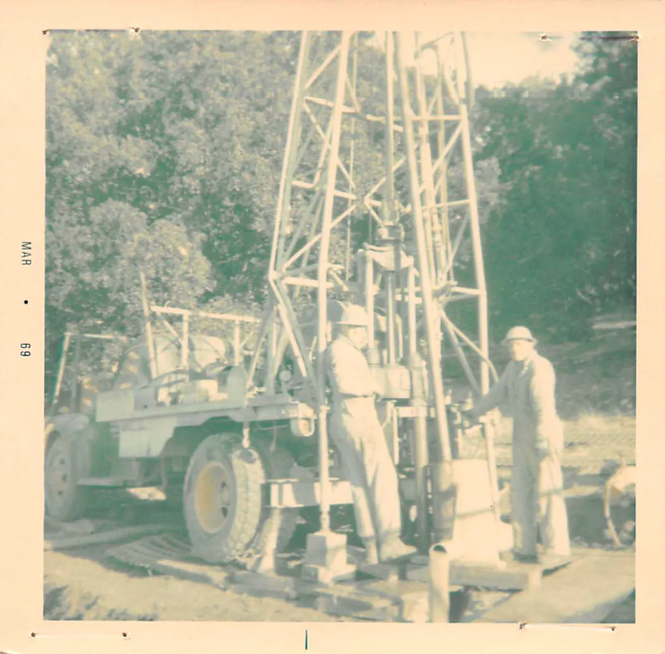 Two workers wearing protective gear operate a drilling rig in an outdoor setting. The rig is mounted on a wheeled vehicle. Trees are visible in the background.
