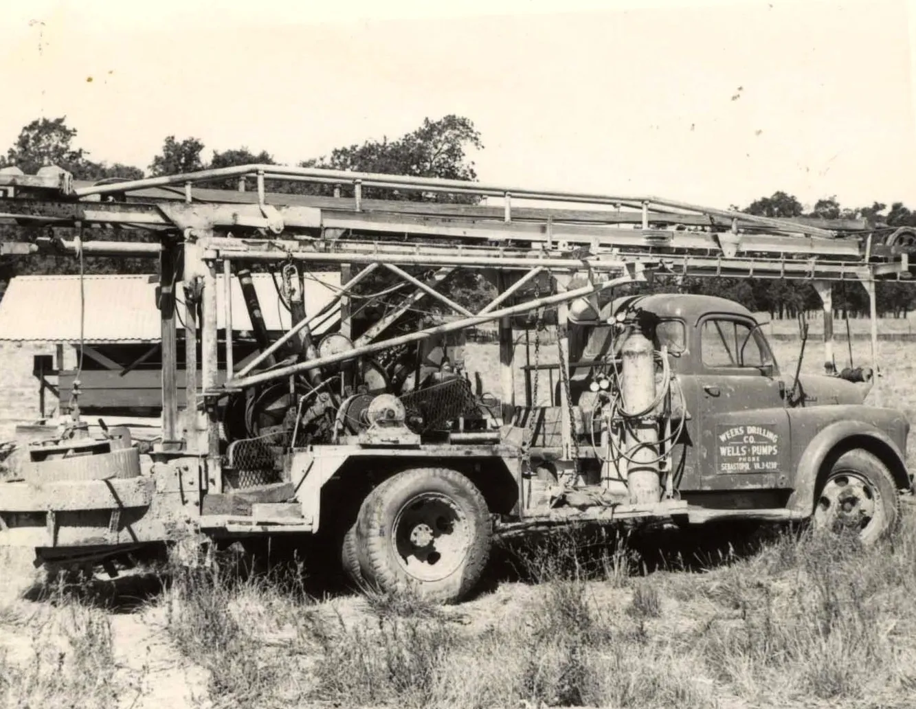  A vintage drill truck parked in a field, with equipment for wells and pumps. The truck bears the Weeks Drilling & Pump Company logo and is surrounded by grassy terrain.