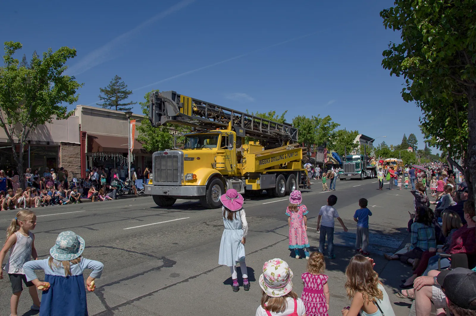 Large yellow drilling truck from Weeks Drilling & Pump Company drives in a parade, surrounded by spectators and children on a sunny day.