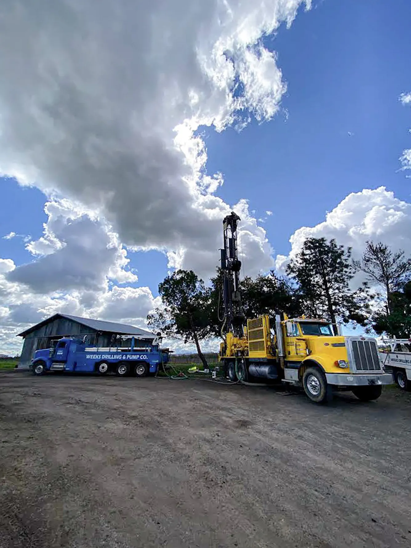  Large drilling equipment beside a blue truck labeled "Weeks Drilling & Pump Co." against a backdrop of cloudy sky and trees.